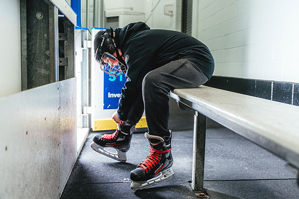 A hockey player sits on a bench in a locker room, leaning forward to tie the laces of their black and red ice skates. They are wearing a black helmet with a protective cage, a dark hoodie, and gray sweatpants. The entrance to the ice rink is visible in the background, with bright light shining through.